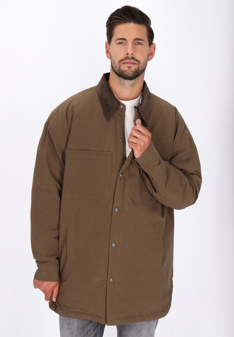 Mann mit Bart, der eine braune, geknöpfte Jacke mit dunklem Kragen und hellhem Hemd trägt, die Jacke anpasst, neutraler Hintergrund.