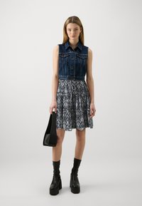 Denim ärmlös jacka med knappar fram, tillsammans med en lager-på-lager svartvit mönstrad kjol. Modellen håller en svart axelväska.