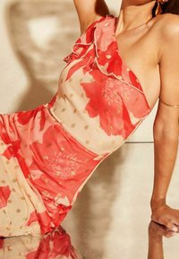Ein ärmelloses Kleid aus transparenter Stoff mit rotem Blumenmuster und goldenen Punkten. Mit Rüschen am Ausschnitt und drapierten Details.