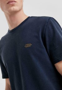 Man in een donkerblauwe T-shirt met kleine gele tekst op de borst die "NEEM EEN GOED VERDIEND PAUZE" leest, tegen een effen achtergrond.