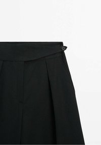Zwarte hoge taillebroek met plooien, voorzien van een stoffen riem en een knoopsluiting. Inclusief zijzakken en een gladde textuur.