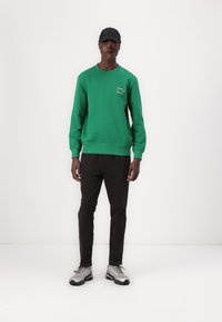 Felpa verde con polsini e orlo a coste, logo bianco sul petto. Abbinata a pantaloni neri e scarpe sportive argento. Cappellino nero indossato.
