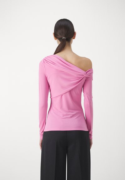 Haut rose à manches longues avec un design asymétrique dénudé d'une épaule, des détails de tissu drapé et une silhouette ajustée. Associé à un pantalon noir à jambes larges.
