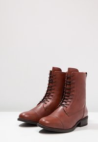 Botas de cuero marrón hasta el tobillo con un diseño de cordones y punta redonda. Presentan una textura suave y un tacón bajo apilado.