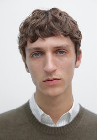 Portret van een jonge man met kort, krullend bruin haar, in een licht gekeerd overhemd onder een donkergroene trui. Neutrale achtergrond.