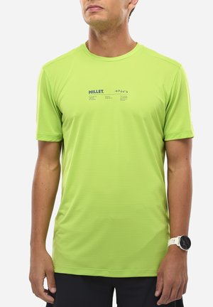 Homme portant un maillot de sport vert citron à manches courtes avec le logo "MILLET" et un short noir, montre de sport blanche au poignet gauche, debout devant un fond blanc.