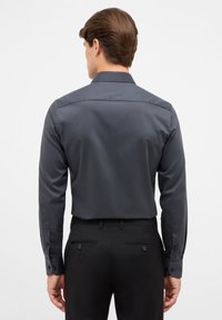 Maglietta grigia a maniche lunghe con colletto strutturato, texture liscia e polsini con bottoni, abbinata a pantaloni neri con tasche posteriori e dettagli con bottoni.