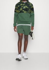 Hoodie verde tie-dye con logo, abbinato a pantaloni corti coordinati. Canottiera bianca e sneakers grigie. Tiene una banda elastica.