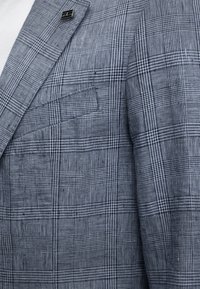 Κοντινή λήψη ενός μπλε και γκρι καρό σακακιού με τσέπη στο στήθος και μικρό ορθογώνιο μεταλλικό καρφί στο πέτο.