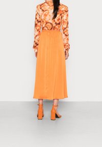 Langes oranges Kleid mit einem langärmeligen Oberteil im Batikmuster, elastischer Taille und glatter Textur; kombiniert mit orangen Absatzsandalen.