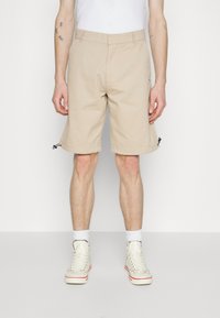 Shorts beige arrivant au genou en tissu léger, avec des poches latérales et des cordons de serrage réglables à l'ourlet. Portés avec des baskets blanches.