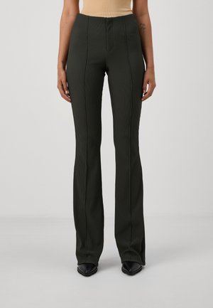 Personne portant un pantalon noir côtelé taille haute à jambes droites et des chaussures pointues noires, debout devant un fond uni gris clair.