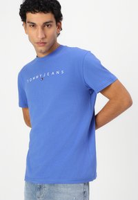 Mėlynas medvilninis T-shirt su trumpomis rankovėmis ir apvaliu kaklu, priklausantis su balta "TOMMYJEANS" tekstu ir mažo logotipo akcentu priekyje.