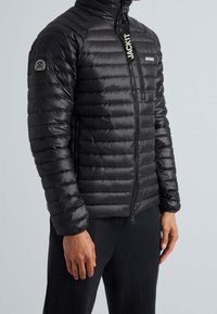 Veste matelassée noire avec col montant, design matelassé horizontal, fermeture éclair frontale et logo sur la poitrine et la manche. Texture lisse.