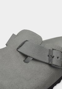 Nærbillede af grå ruskindssko med justerbar rem og metalspænde på hvid baggrund.