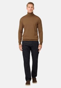 Rjav pulover s turtleneck ovratnikom, izdelan iz pletenine, v kombinaciji s temnimi kavbojkami in rjavimi semiš čevlji. Model stoji z rokami ob straneh na beli podlagi.