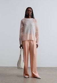 Hellrosa und cremefarbener, gestrickter Pullover mit weitem Ausschnitt, kombiniert mit weichen rosa Hosen. Das Modell hält eine weiße Handtasche. Glatte Texturen sind sichtbar.