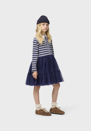 Pige iført navy hue, stribet langærmet top, navy tylskørt, hvide blonde sokker og brune sneakers, stående mod hvid baggrund.