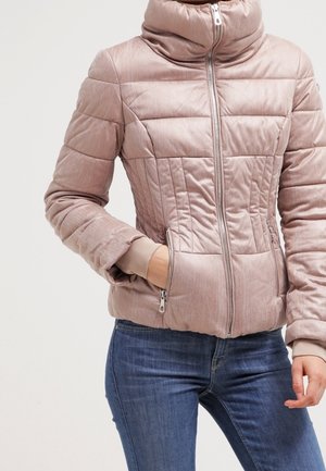 Kvinde iført en lyserød, quiltet jakke med høj krave, lynlås og lynlåslommer, kombineret med blå jeans.