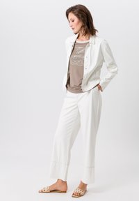 Witte jas, bruine met patronen bedekte top, en witte wijde broek. Beige sandalen met decoratieve uitsparingen. Glad textuur en minimalistisch ontwerp.