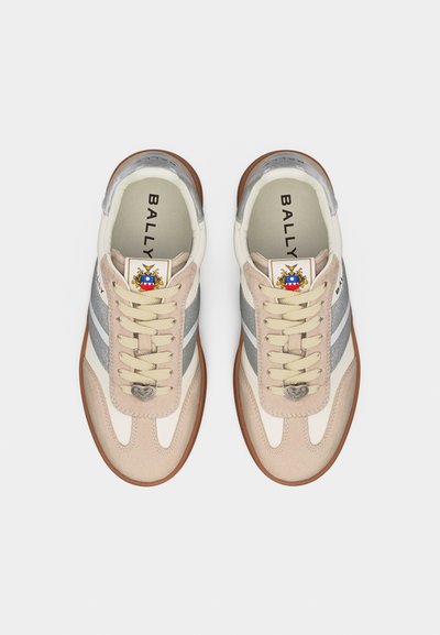 Paire de baskets Bally beige, blanches et argentées avec lacets, breloques en forme de cœur sur les lacets, et un blason coloré sur la languette, vues de dessus.