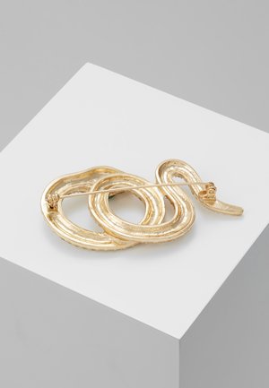 Broche de couleur dorée en forme de serpent enroulé avec une fermeture à épingle, exposée sur un socle blanc sur fond gris.