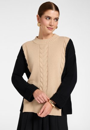 Vrouw met gouden oorbellen en armband, gekleed in een beige en zwarte kabelgebreide trui, handen samengevouwen voor, kijkt opzij.