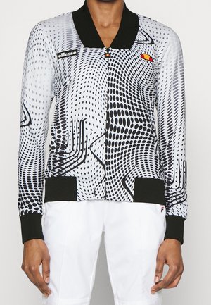 Czarna i biała kurtka na zamek z długim rękawem, z wzorem w kropki, czarnym kołnierzem i mankietami, noszona z białymi spodniami, z naszywkami z logo na piersi i kieszeniach.