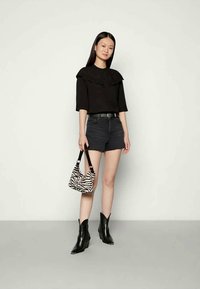 Mulher jovem vestindo blusa preta com babados, calções de denim pretos, botas pretas de tornozelo, segurando uma mala com padrão de zebra, de pé contra um fundo claro e liso.