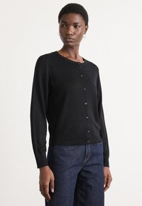 Svart cardigan laget av mykt materiale, med rund hals, knappestolpe foran og ribbestrikket kant. Sammen med mørkeblå jeans.