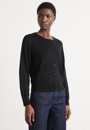 Svart cardigan laget av mykt materiale, med rund hals, knappestolpe foran og ribbestrikket kant. Sammen med mørkeblå jeans.
