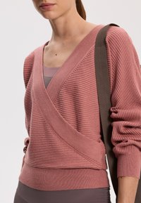 Ribbed knit sweater in soft rose, featuring a crossover design, voluminous sleeves, and a detailed hem. Paired with a dark strap.