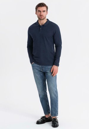 Marinefarbener Langarm-Polohemd aus Baumwolle, mit einer Drei-Knopf-Leiste, kombiniert mit hellblauen Jeans und schwarzen Schnürschuhen. Einfaches Design.