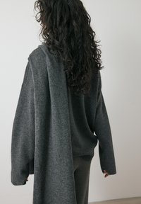 Grijze gebreide trui met een losse pasvorm en lange mouwen, met een waterval-stofdetail dat over de rug valt. Zachte textuur en warm materiaal.