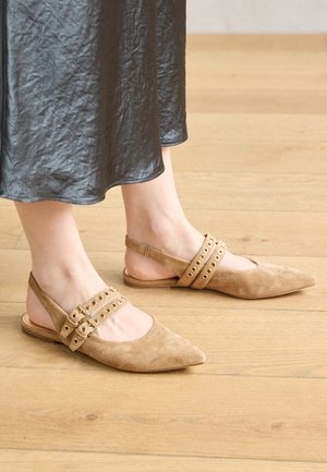 Beige ruskinds slingback-sko med spidse tæer, med to dekorative stropper med metaløjer, båret uden sokker.