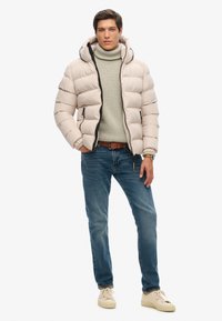 Chaqueta tipo abrigo en beige claro con patrón acolchado horizontal, cremallera negra y capucha. Llevada sobre un suéter de cuello alto gris claro y jeans azules.