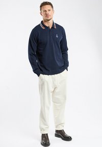 Polo à manches longues bleu marine avec col rayé rouge et blanc, logo sur la poitrine, associé à un pantalon en velours côtelé crème et des bottes noires à la cheville.