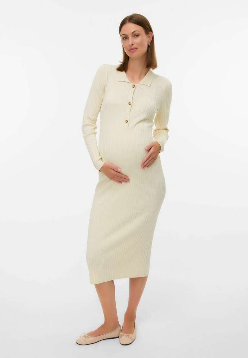 Vestito premaman a coste color crema fino al ginocchio, con maniche lunghe, scollo con colletto e finte decorazioni con bottoni. Indossato con ballerine chiare.