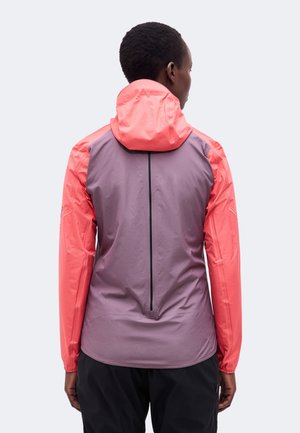 Vue de dos d’une personne présentant une veste à capuche corail et mauve, avec une longue fermeture éclair noire dans le dos et un pantalon noir.