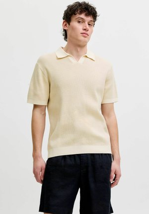 Ung mand med krøllet hår iført en cremefarvet strikket kortærmet polo shirt og sorte shorts, stående mod en ensfarvet baggrund.