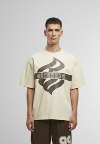T-shirt oversize beige avec un design graphique sombre et le mot "ROCAWEAR" centré, fabriqué en coton doux.