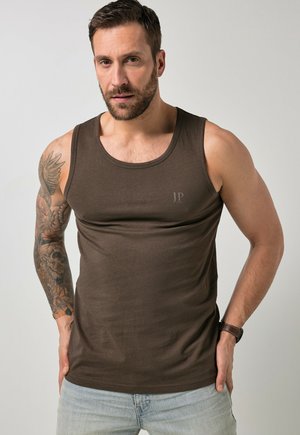 Mann mit Bart, der ein braunes ärmelloses Hemd und hellblaue Jeans trägt, zeigt einen Tattoo-Ärmel am linken Arm und eine Armbanduhr am rechten Handgelenk.