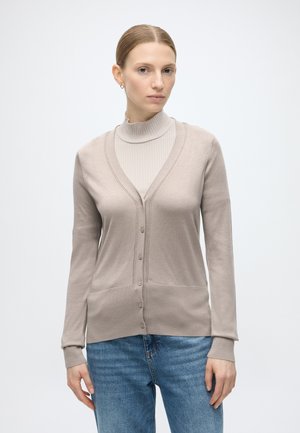 Femme portant un cardigan beige boutonné sur un pull col roulé beige clair et un jean bleu, debout devant un fond blanc uni.