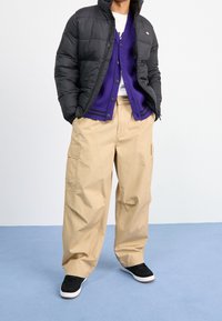 Persona con giacca a vento nera, cardigan viola abbottonato, pantaloni cargo beige e sneakers nere che si trova su un pavimento blu contro uno sfondo bianco.