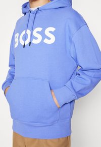 Ljusblå hoodie med en framficka, vit "BOSS" logotyp, justerbar dragsko på huven och ribbade ärmslut samt nederkant.