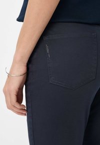 Donkerblauwe slim-fit broek met een achterzak die een subtiel ontwerp en contrasterende stiksels heeft. De hand rust lichtjes op de heup.