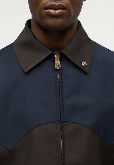 Κοντινό πλάνο ενός ατόμου που φοράει μπλε ναυτικό και μαύρο μπουφάν με χρυστό φερμουάρ και χρυσό κουμπί στο γιακά, με ορατά τα χείλη και το κάτω μέρος του προσώπου.