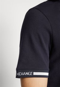 T-shirt polo noir en tissu texturé, avec une bande contrastante sur la manche marquée "EXCHANGE" en blanc, mettant en valeur le col et le détail des coutures.