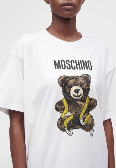 Άτομο φοράει λευκό μπλουζάκι Moschino με εικόνα αρκουδάκι τυλιγμένο σε κίτρινη και λευκή μεζούρα μπροστά.