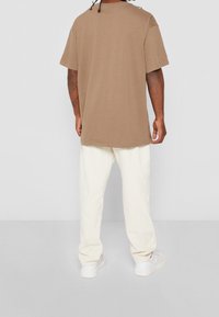 Hnědé oversized tričko s krátkým rukávem, v kombinaci s krémovými volně střiženými kalhotami a bílými teniskami, pohled zezadu.